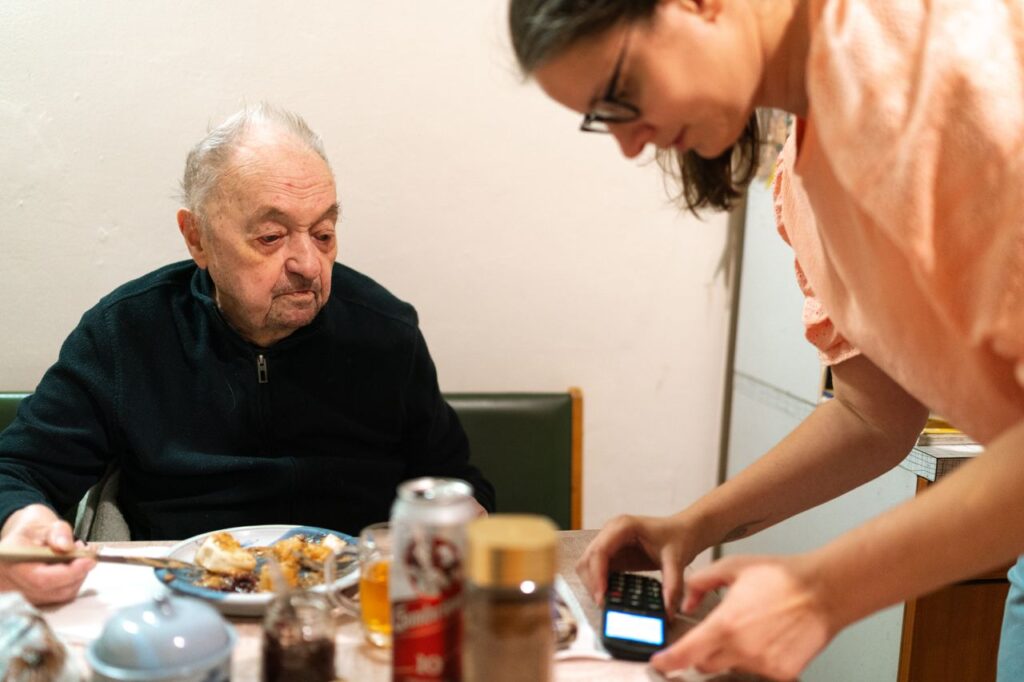 A caregiver assists an elderly man during mealtime at an indoor setting. Captured in Praha.