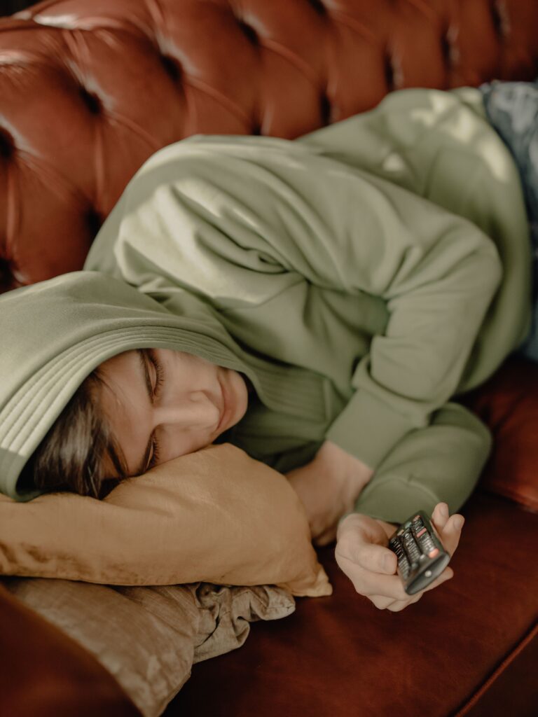 Young man in green hoodie resting on leather sofa holding remote control, cozy home setting.