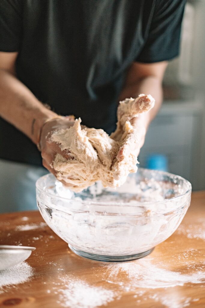 Hands kneading bread dough in a glass bowl on a wooden table for homemade cooking.
