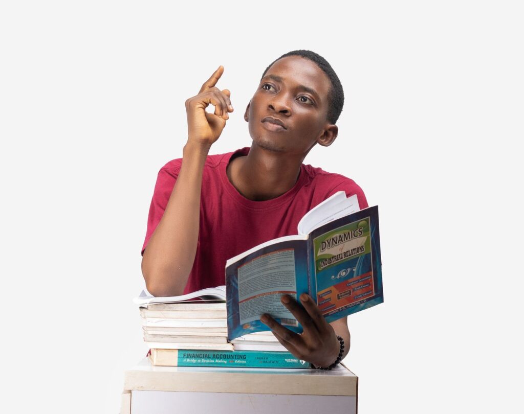 A young man in a studio setting, deep in thought with a stack of books and a white background.