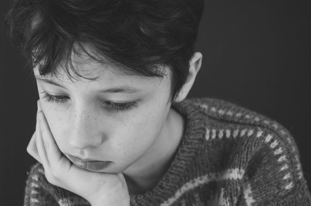 Black and white of sad boy with short dark hair in casual clothes covering face with hand while looking down
