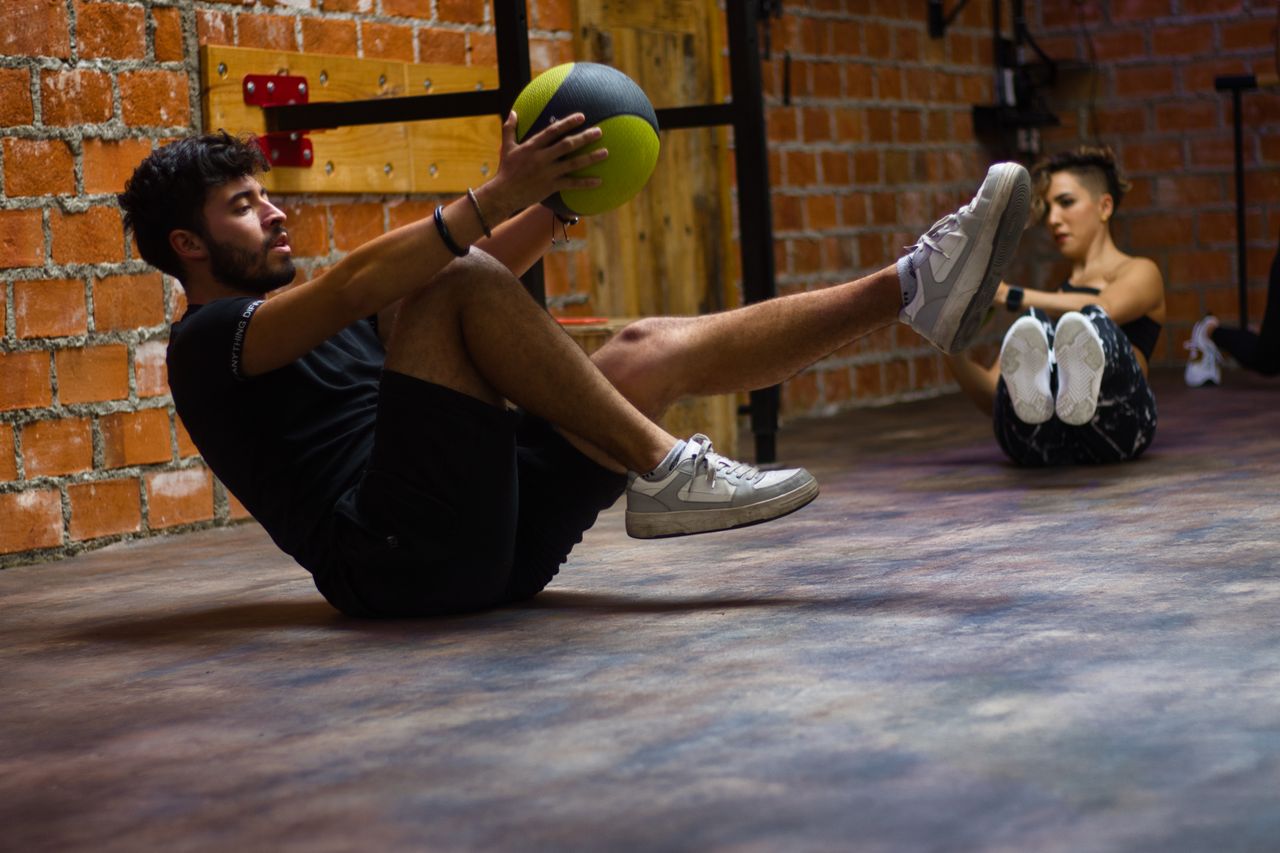 A man and woman perform core exercises with a medicine ball in a brick wall gym.