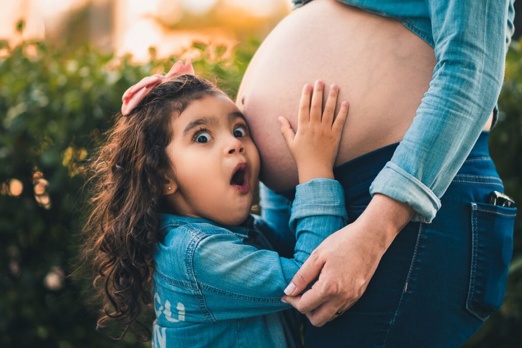 A young girl joyfully embraces her pregnant mother's belly in a warm outdoor setting.