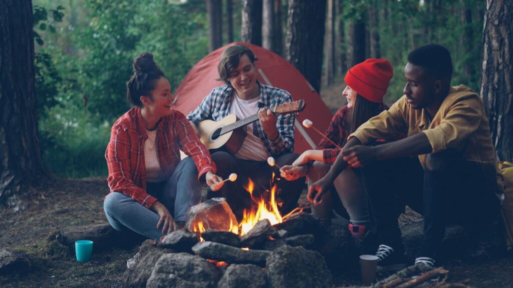 Four friends around a campfire, roasting marshmallows and playing guitar outdoors.