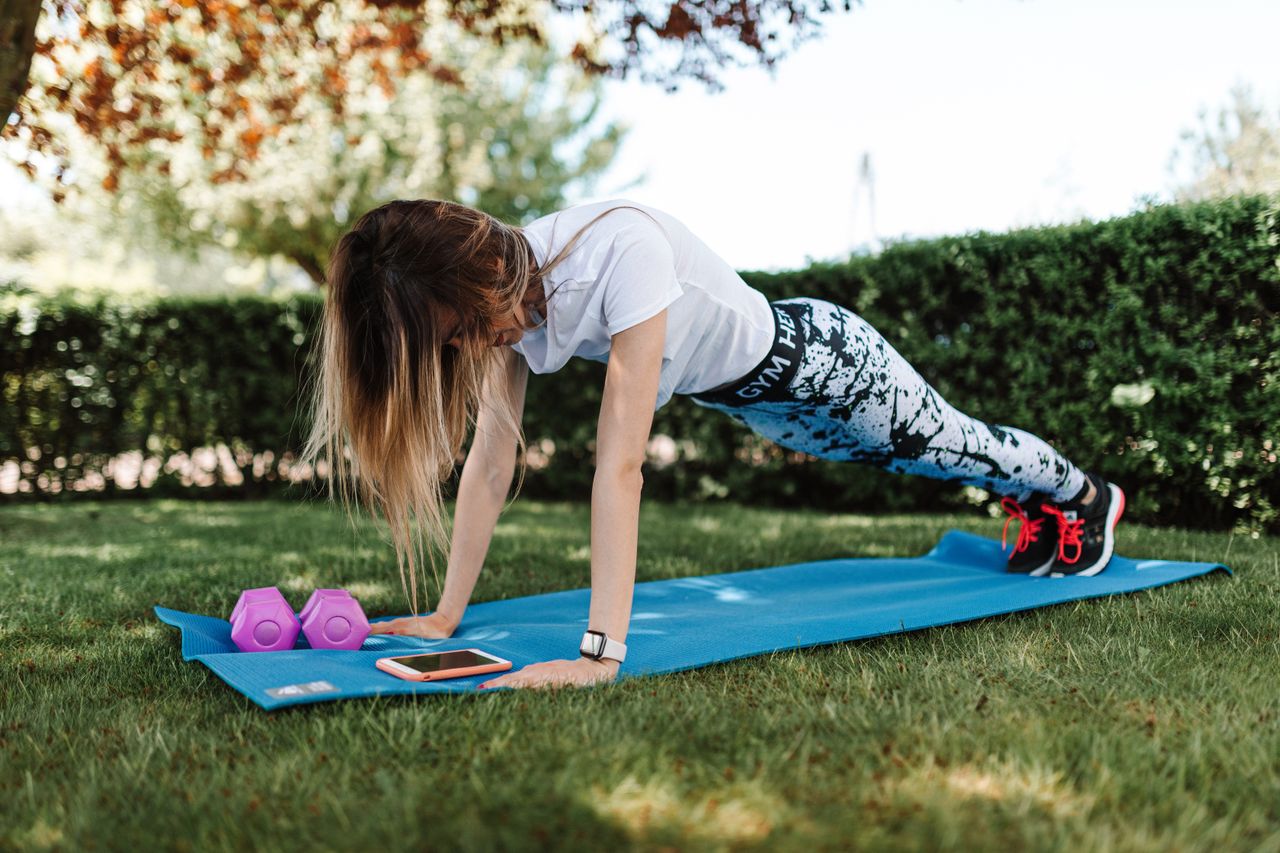 Woman doing push-ups on a yoga mat outside with dumbbells and a smartphone nearby.