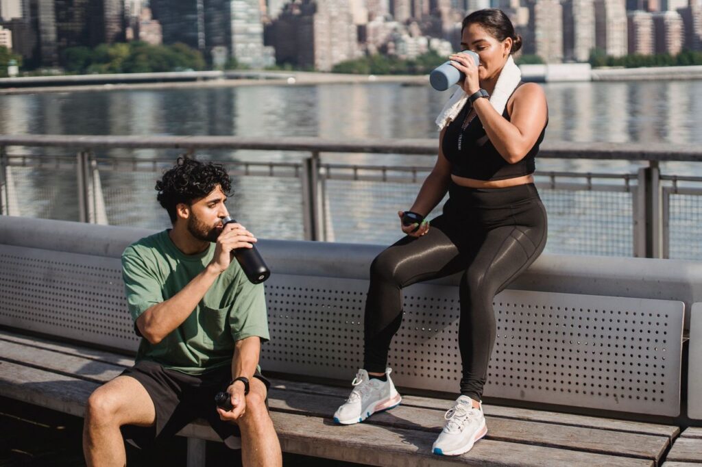 A young couple takes a break by the waterfront to hydrate after exercising outdoors in a sunny city setting.